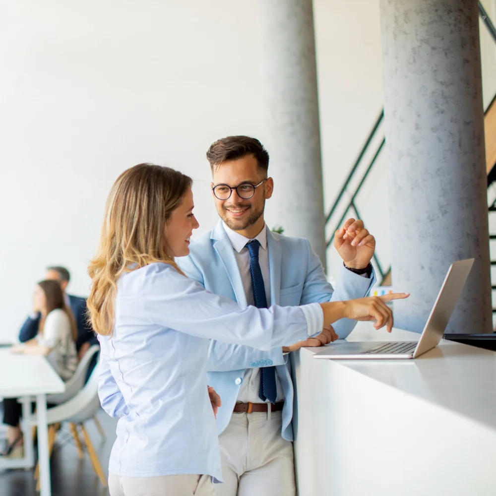 Zwei junge Business-Professionals besprechen gemeinsam ein Projekt an einem Laptop, im Hintergrund ein modernes Büro mit weiteren Kollegen