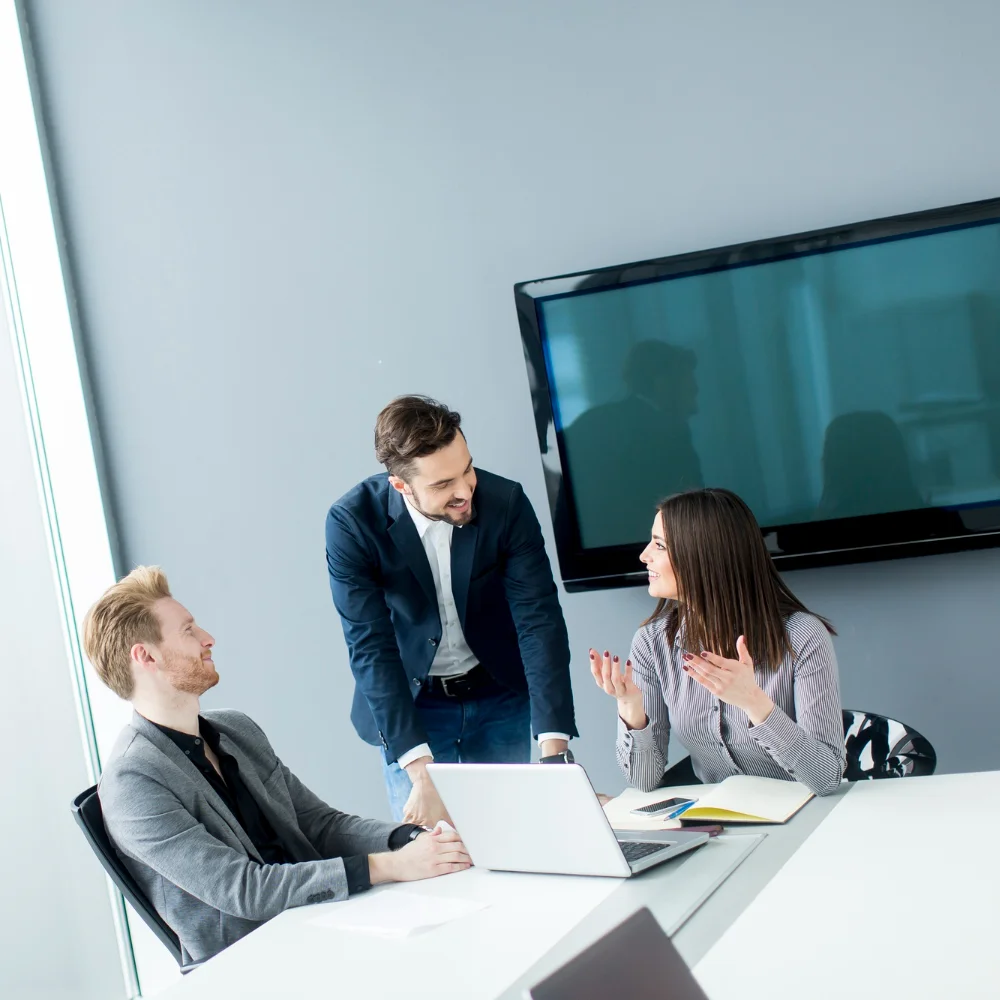 Drei Personen in einem modernen Konferenzraum im Gespräch, mit Laptop und Notizen auf dem Tisch
