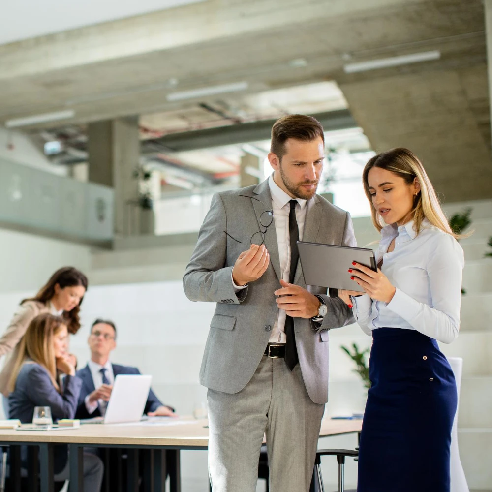 Zwei Geschäftspersonen im Gespräch vor einem Tablet in einem modernen Büro, im Hintergrund ein Team in einer Besprechung