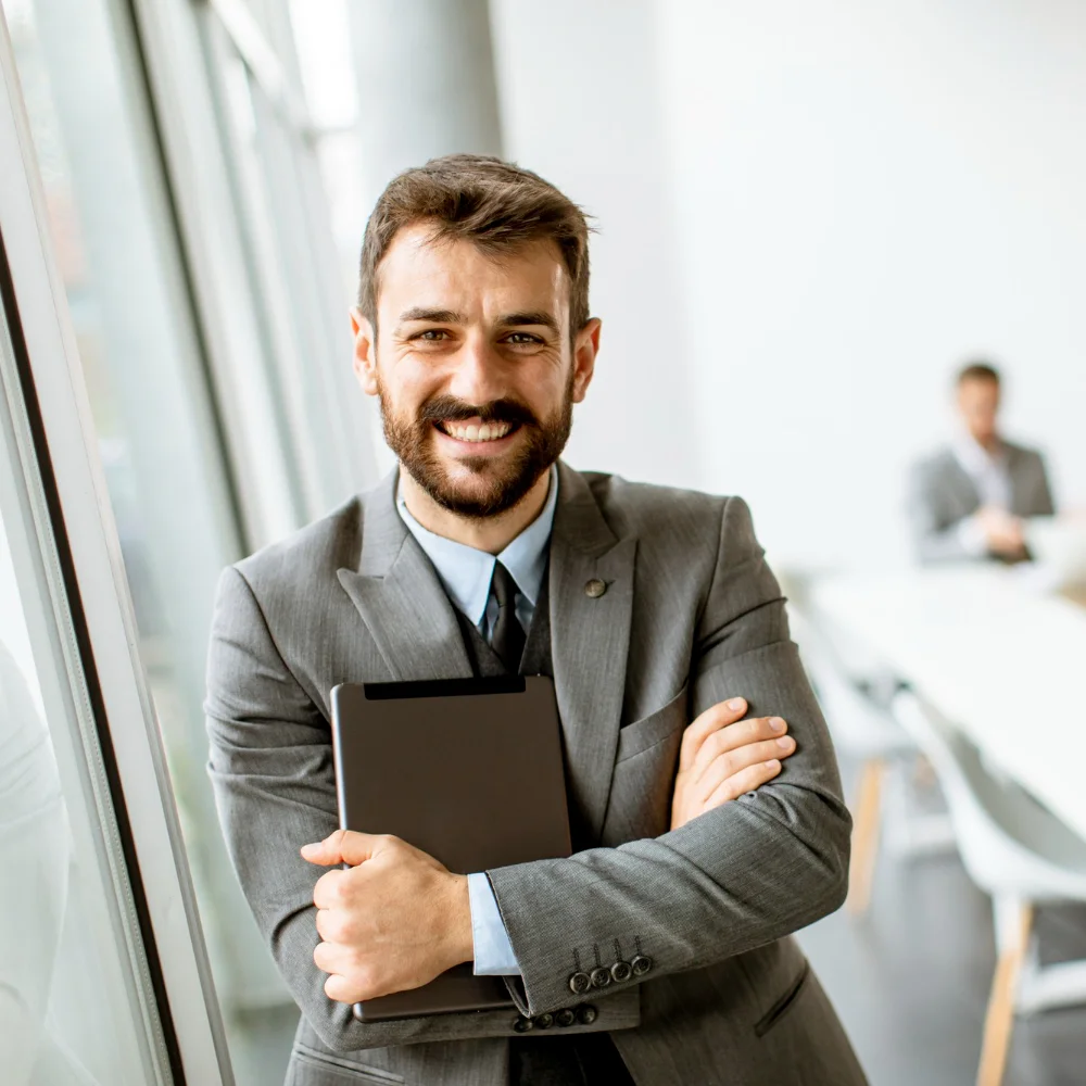 Ein lächelnder Geschäftsmann im Anzug steht mit einem Tablet im Arm vor einem hellen Konferenzraum