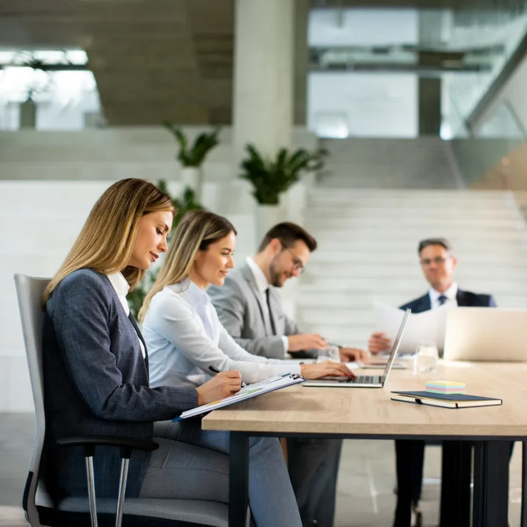 Vier Businesspersonen sitzen konzentriert an einem modernen Besprechungstisch, arbeiten mit Laptop, Notizblock und Unterlagen.