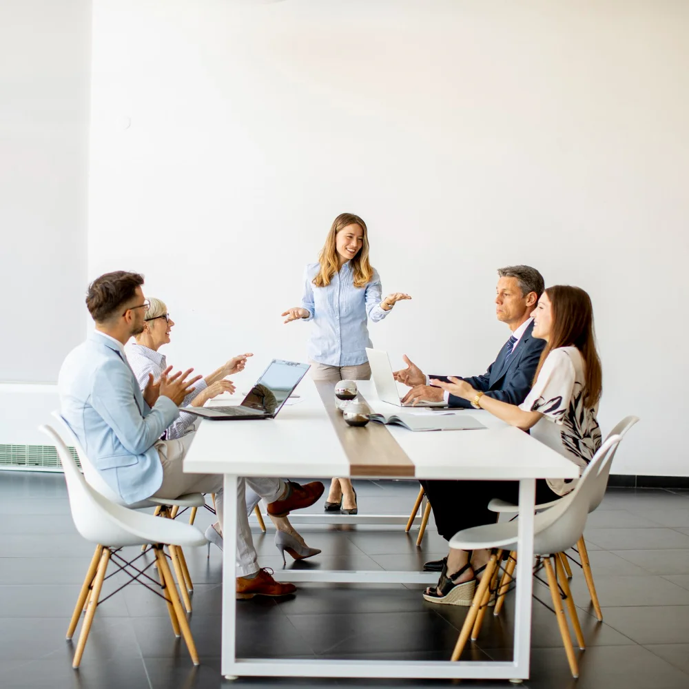 Eine Frau steht vor einem Konferenztisch und spricht mit fünf Kolleginnen und Kollegen, die an Laptops arbeiten und aufmerksam zuhören.