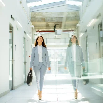 Junge Geschäftsfrau im Anzug geht selbstbewusst durch einen hellen Büroflur, mit Spiegelung an der Glaswand.