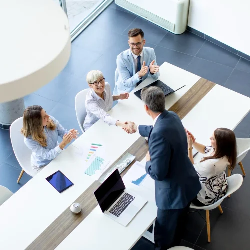 Führungskraft in Businesskleidung gibt einer älteren Kollegin am Konferenztisch die Hand, während das Team applaudiert